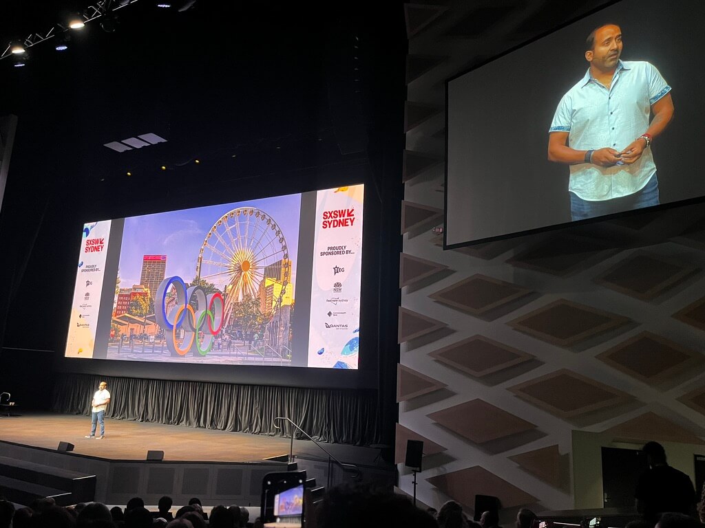 Rohit Bhargava speaking before a packed auditorium, in front of a projected image of the Olympic logo. He is also magnified on a screen to the right of the image.